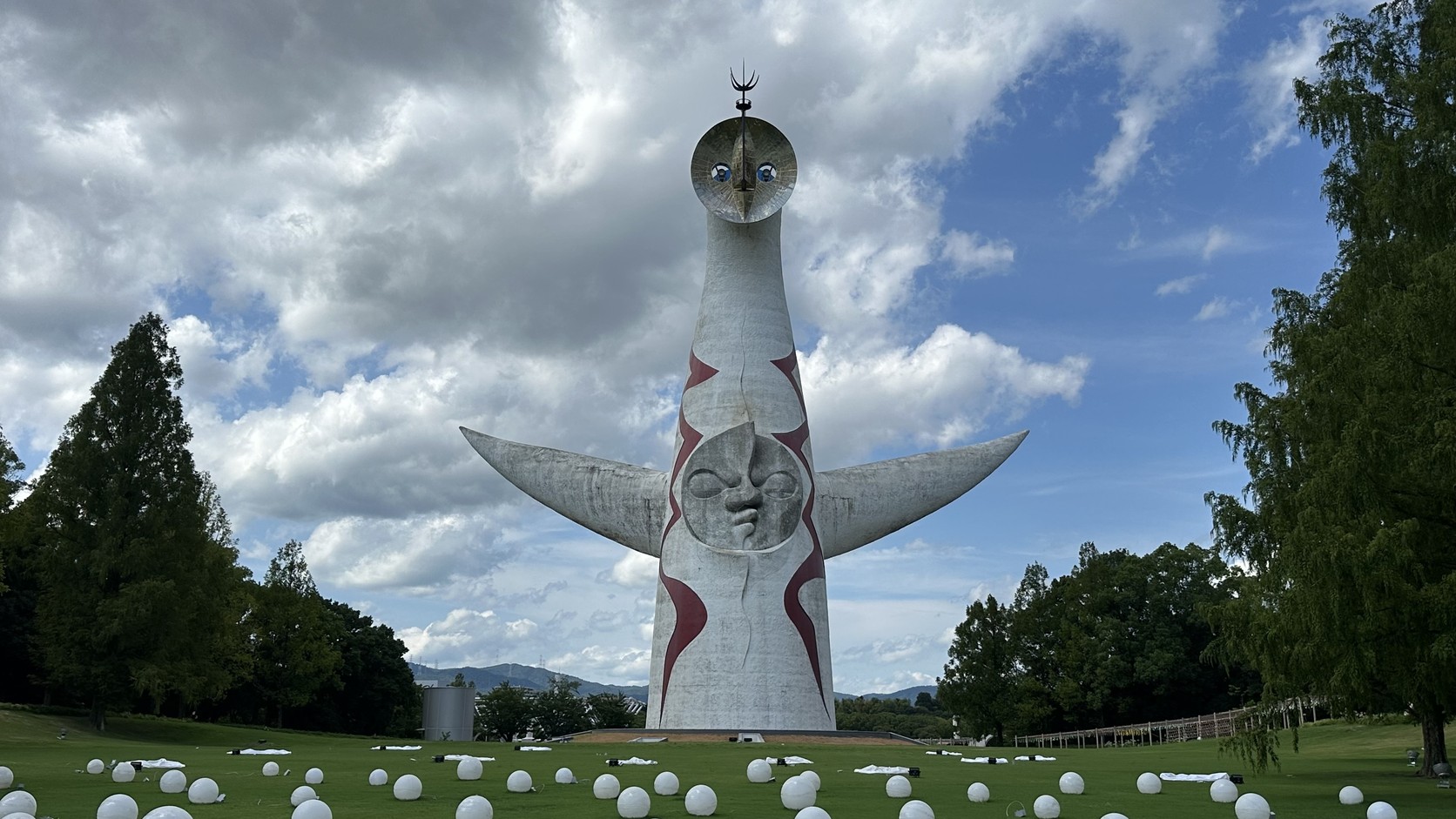 從園內草地望向太陽之塔正面，藍天白雲下的白色塔身與張開的雙臂