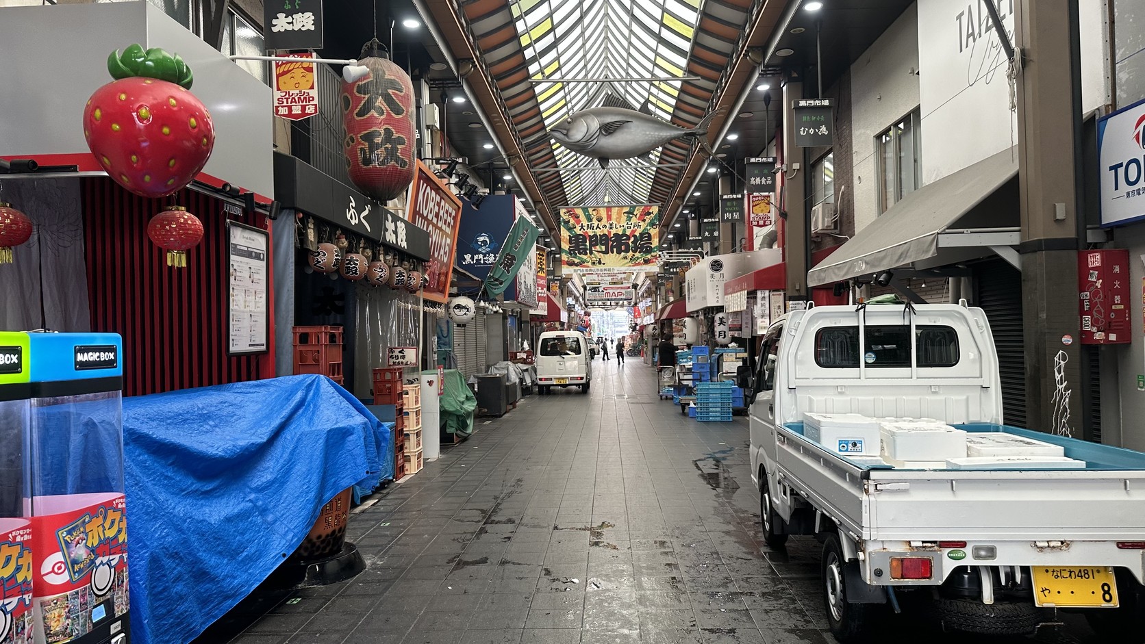 清晨拱廊內的通道，路邊停著小貨車與藍色防水布，沿街許多鐵門尚未拉起