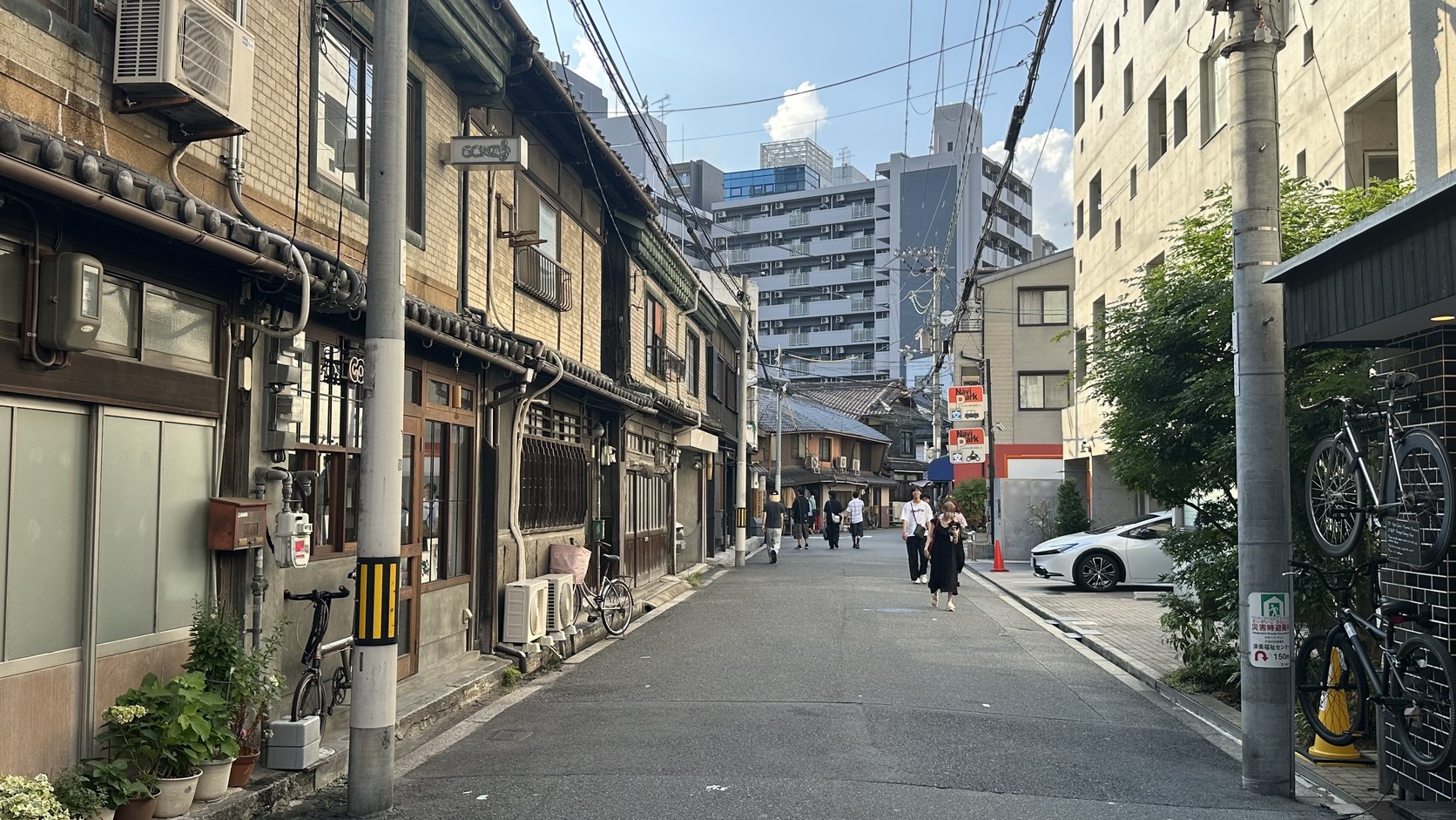 長屋巷弄裡的盆栽與腳踏車，遠方可見現代高樓