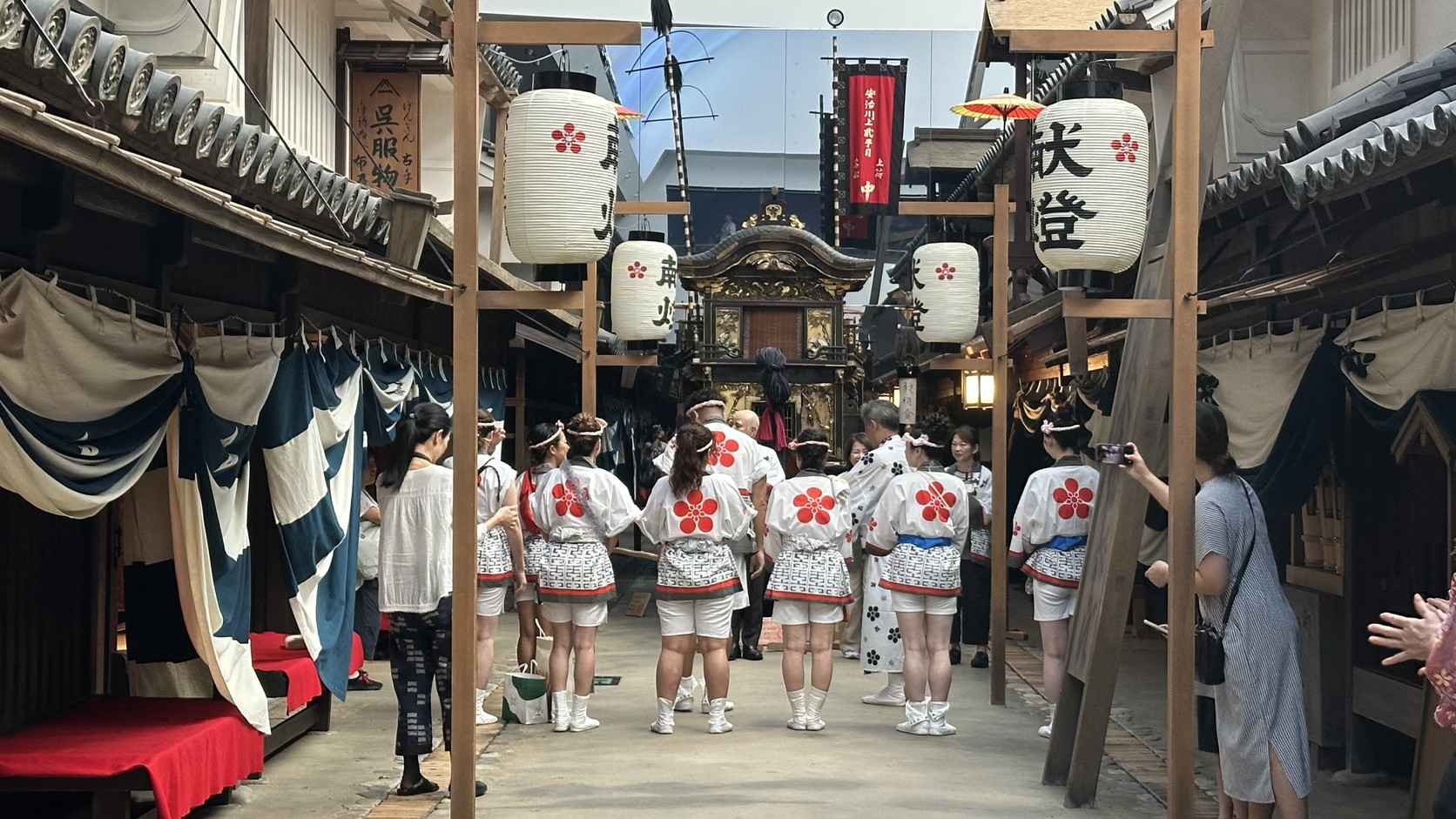 街道中央穿著紅色花紋祭典服的人群背影，後方可見山車