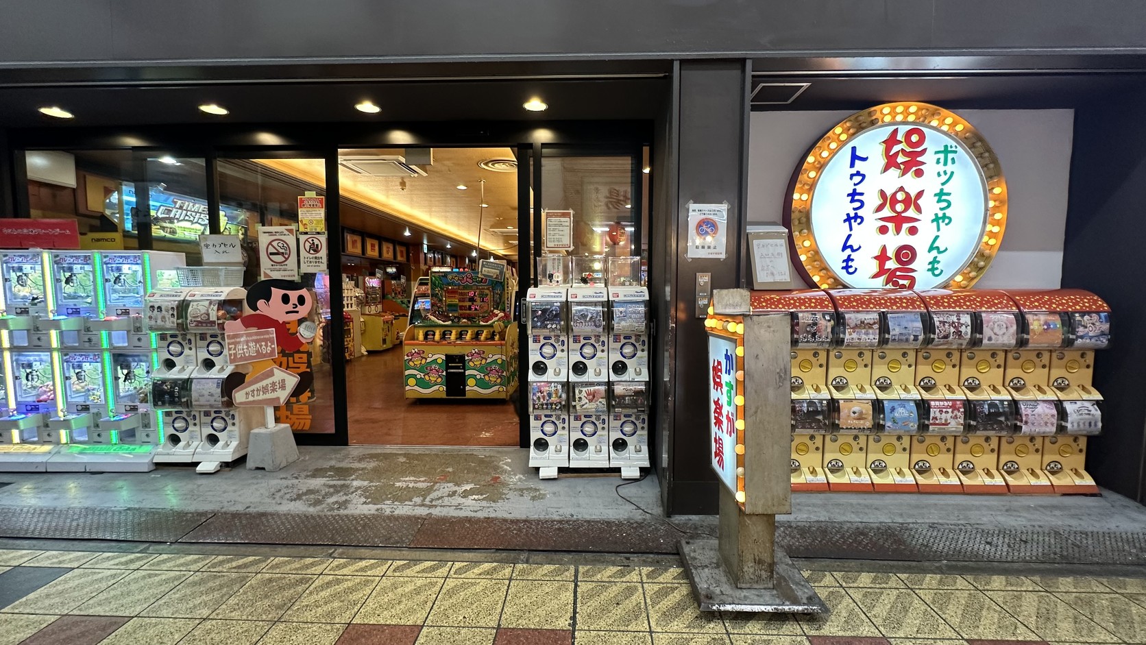 鏘鏘橫丁的かすが娯楽場外觀，門口有燈泡招牌與一排扭蛋機