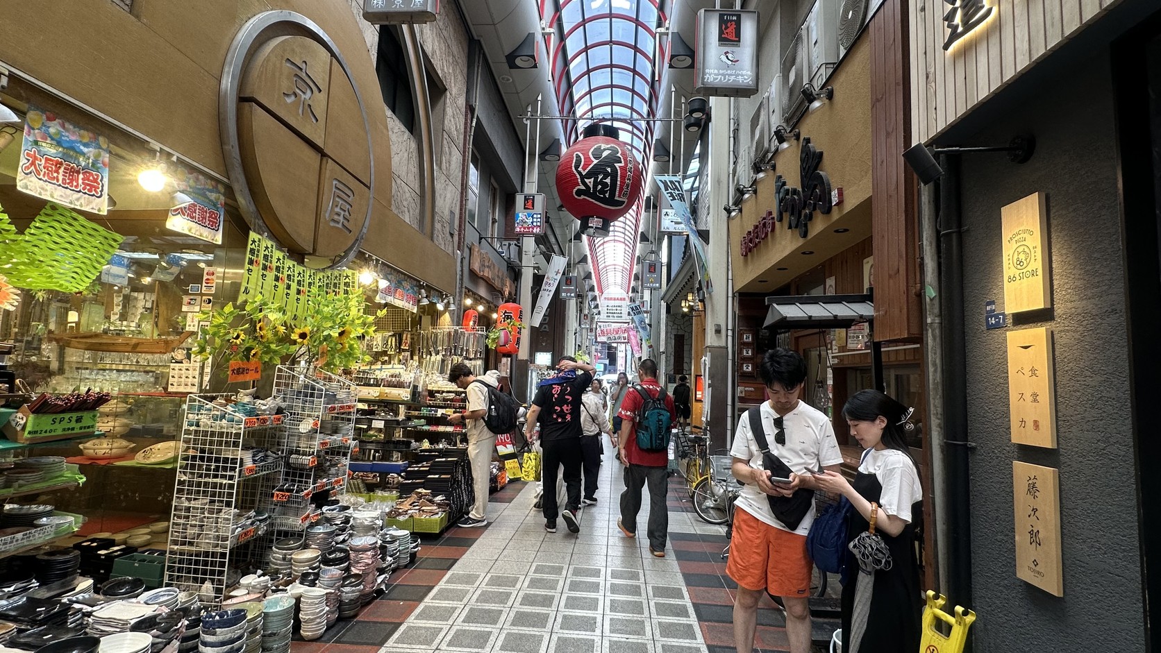 道具屋筋拱廊中懸掛的紅色「道」字燈籠，兩側是堆滿碗盤的食器店與走動的遊客