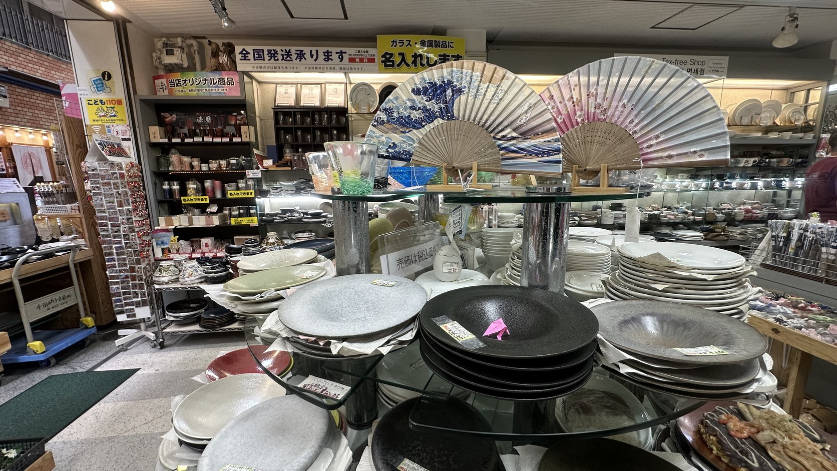 相互商会店內的大型盤子與印有浮世繪風格海浪圖案的摺扇，上方貼著「全国発送承ります」「名入れします」告示