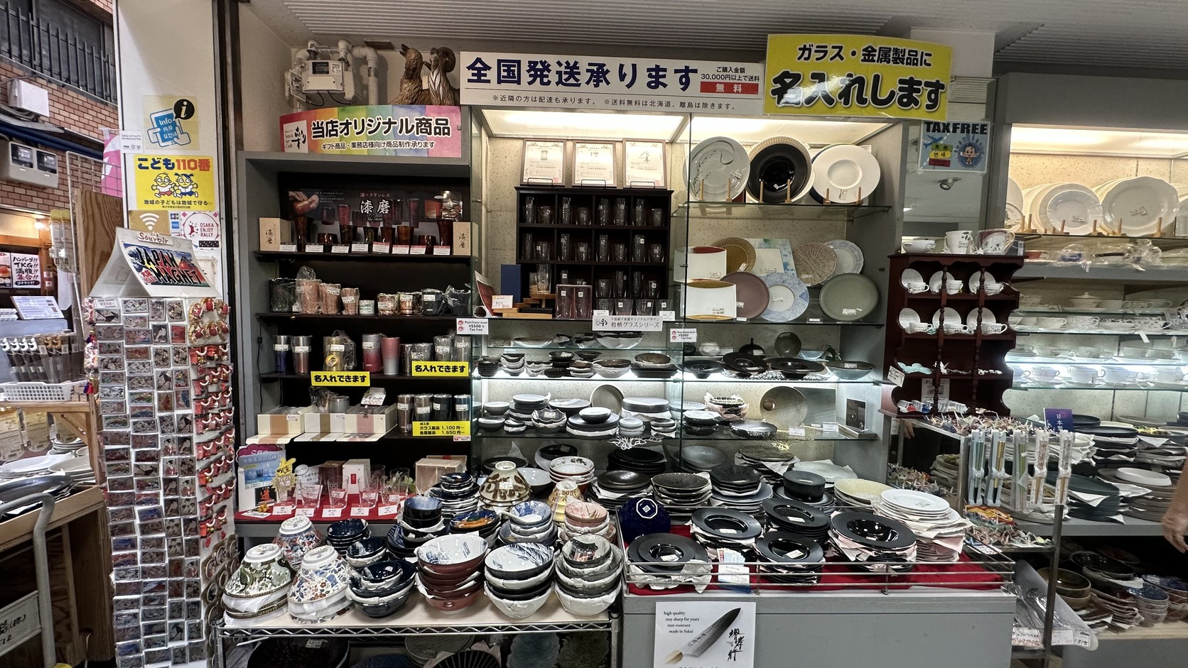 相互商会貨架上密集陳列的碗盤，旁邊貼著「全国発送承ります」「ガラス・金属製品に名入れします」與 TAX FREE 標示