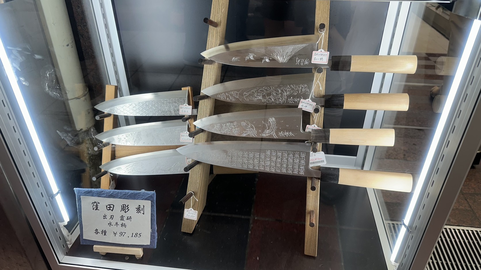 窪田彫刻的出刃刀陳列在玻璃櫥窗裡，價牌標示「各種 ¥97,185」，刀身刻有山形、波浪與細密文字