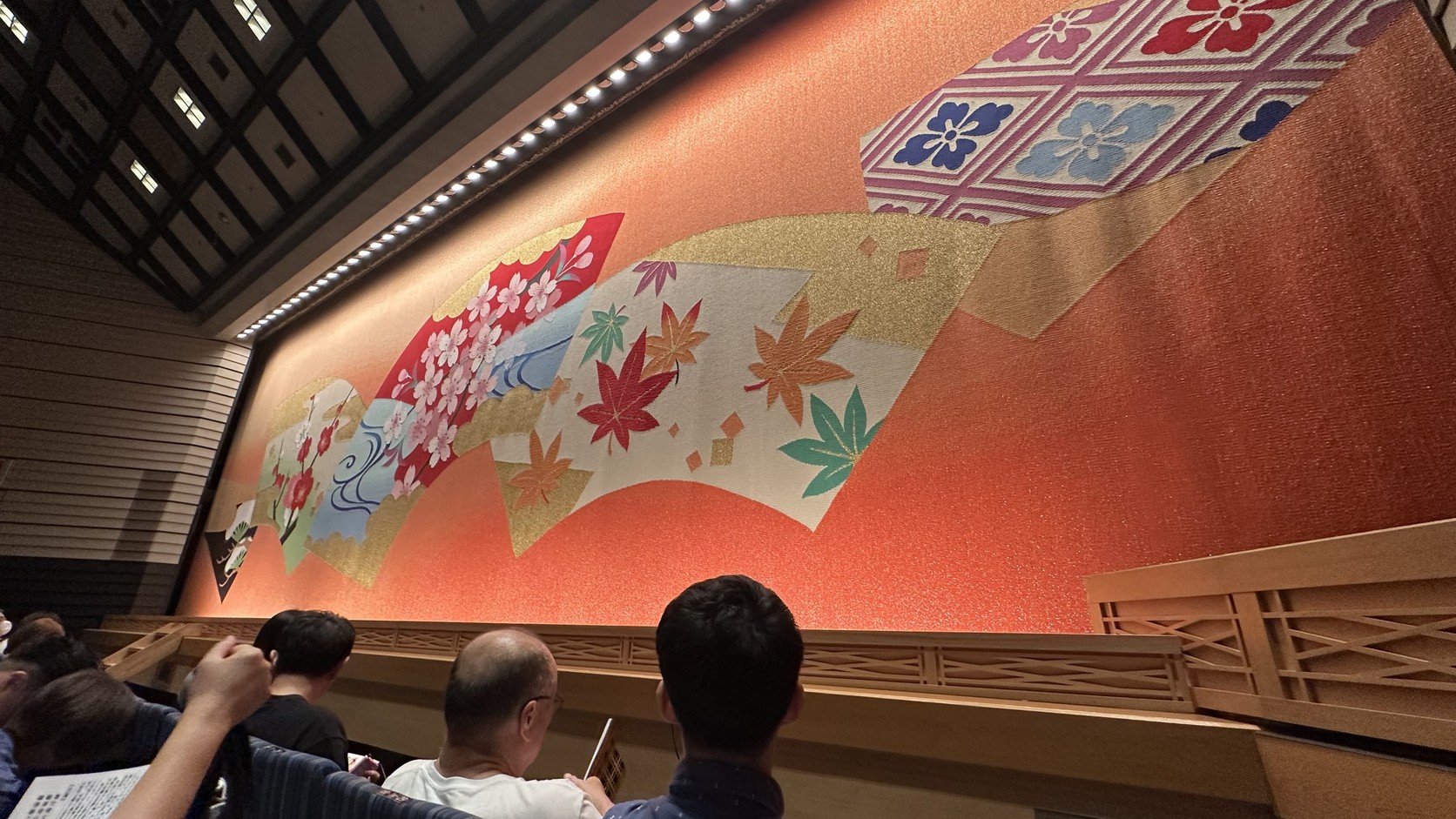 從觀眾席座位望向舞臺大幕，橘色幕面繪有梅、櫻、楓、松與扇面