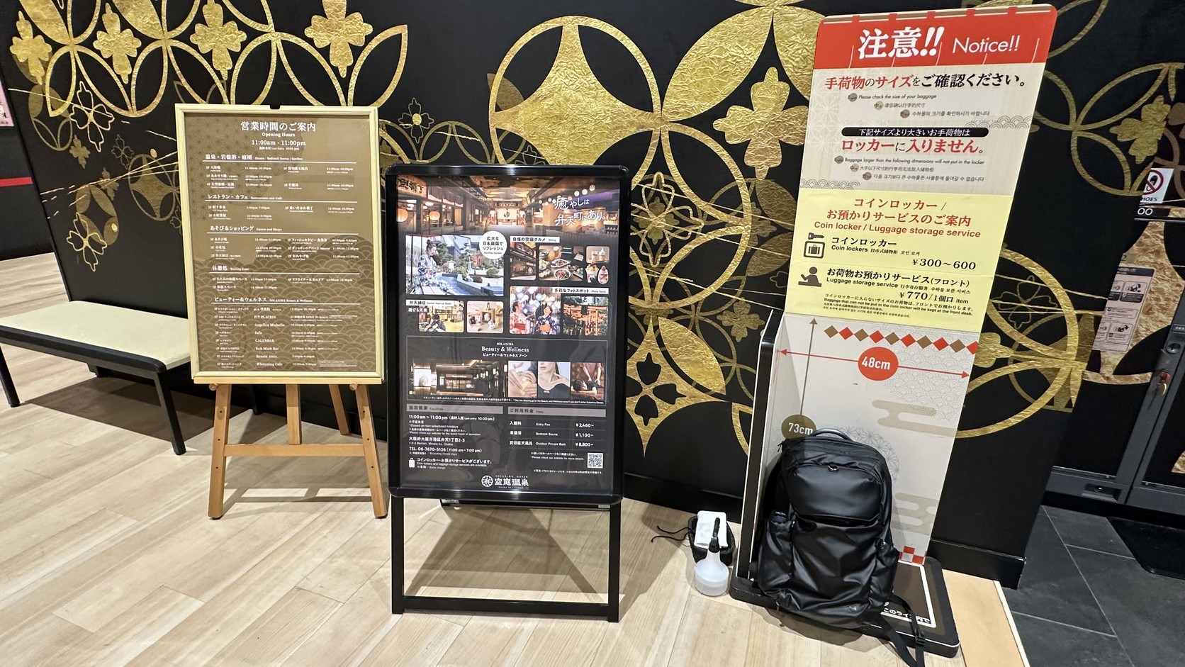 入口旁的行李尺寸告示與黑色後背包，告示寫明寬 48 公分的尺寸參考、投幣置物櫃 300 至 600 日圓與櫃檯寄放每件 770 日圓