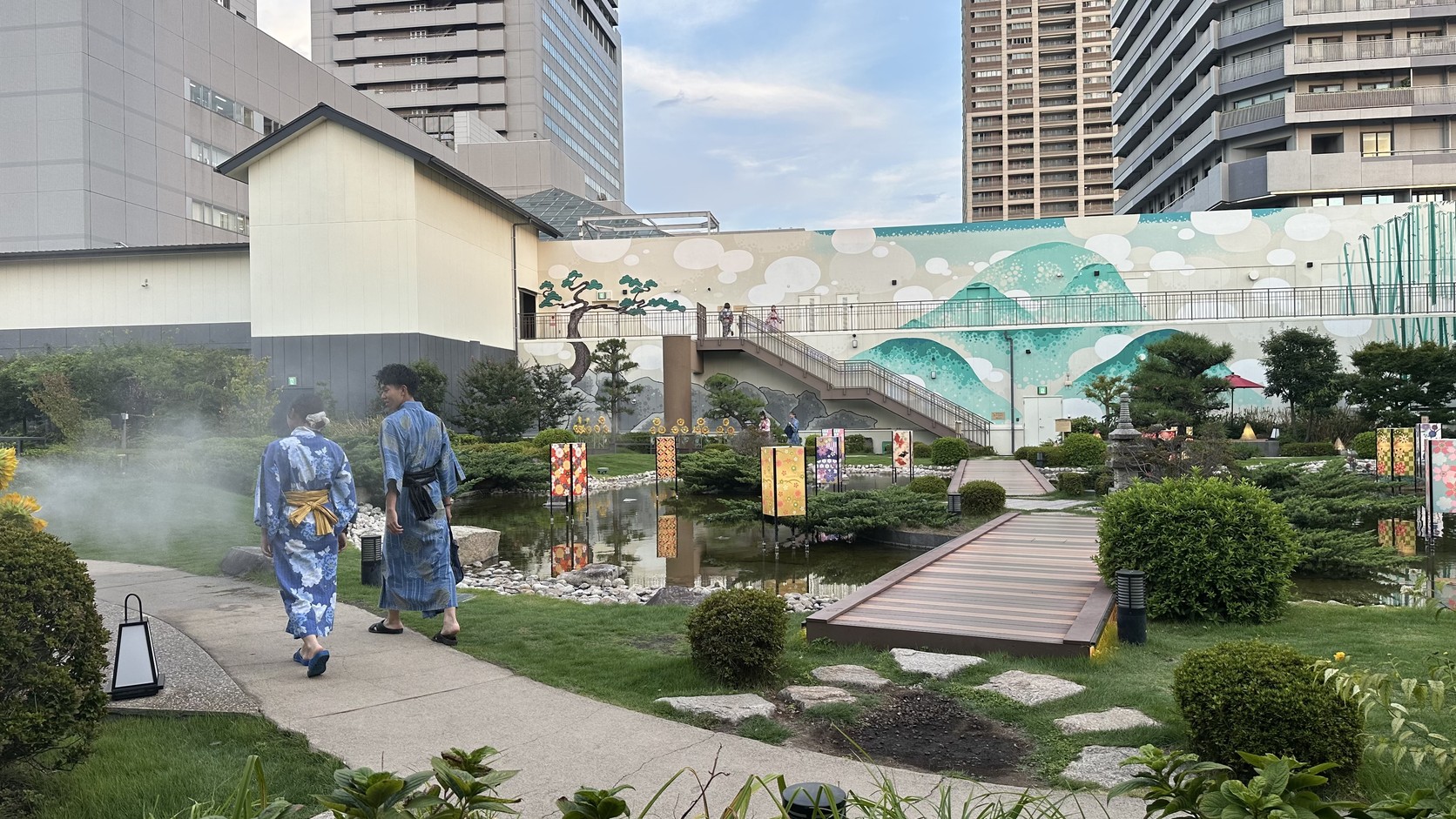 兩位穿浴衣的旅客背影走在天空庭園的步道上，前方是水池與木橋