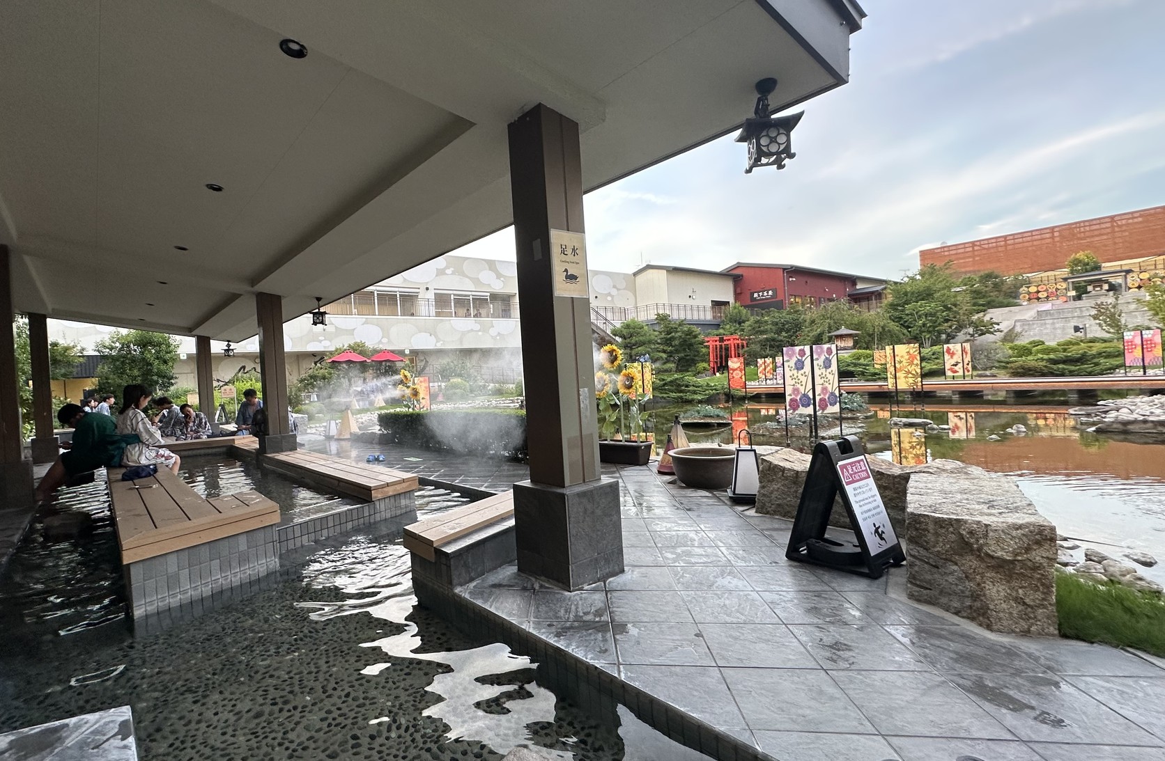 屋簷下的泡腳池，柱子上掛著「足水 Cooling Foot Spa」牌子，旅客坐在池邊泡腳，後方是庭園池塘與燈籠裝飾