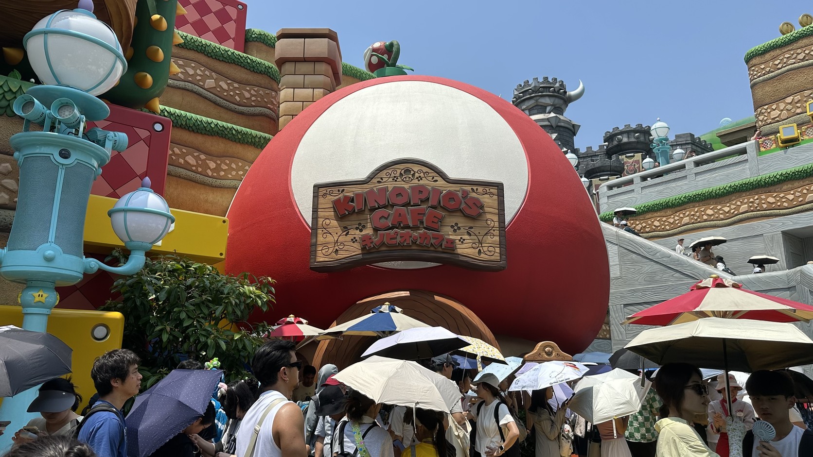 奇諾比奧咖啡店門口大排長龍，撐著陽傘的人群前方是紅底白點的蘑菇屋，招牌寫著 KINOPIO'S CAFE