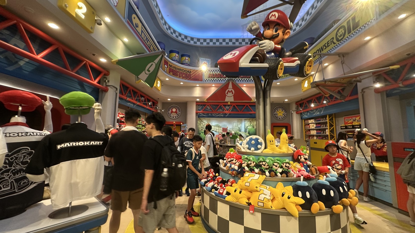 Mario Motors 店內：上方有瑪利歐駕駛的大型賽車模型，四周陳列衣服、帽子、布偶與吊飾
