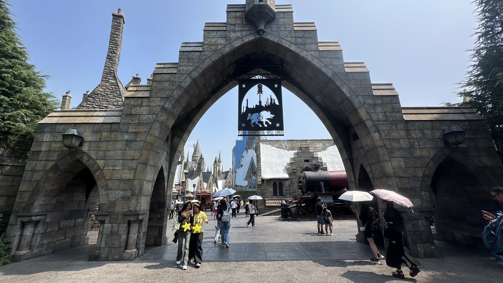 哈利波特魔法世界入口：石造拱門下掛著黑色招牌，拱門後方遠處可見城堡塔樓與停靠的火車，遊客撐著陽傘走進園區