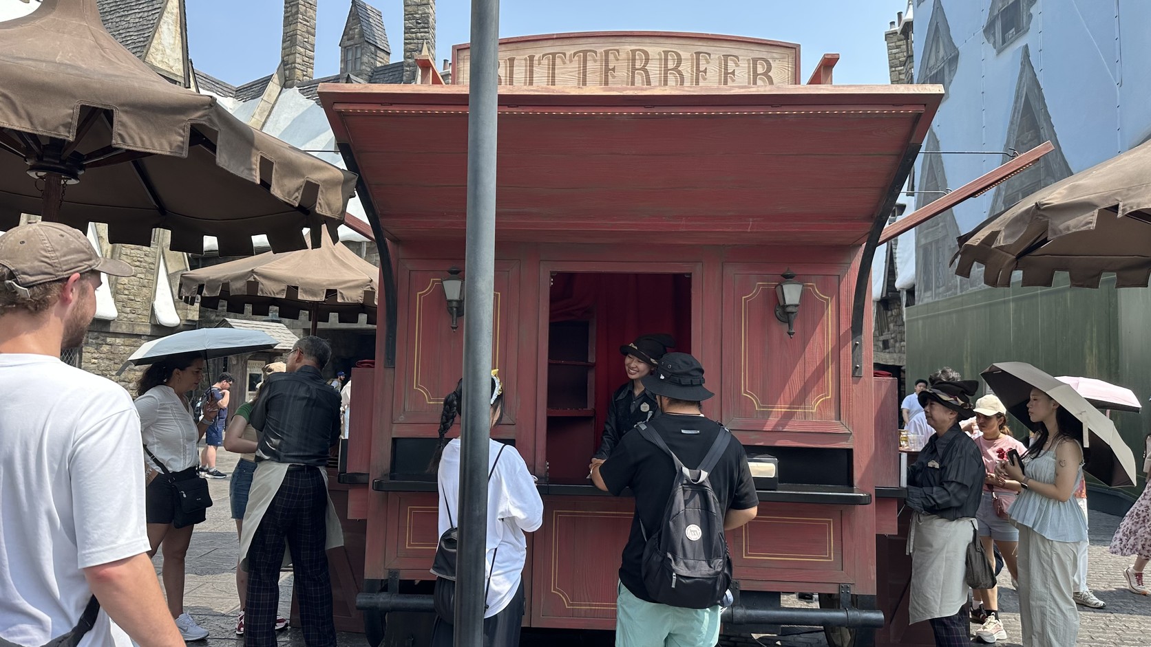 奶油啤酒攤位：紅色木造推車頂端寫著 BUTTERBEER，前方有人排隊，不少遊客撐著陽傘