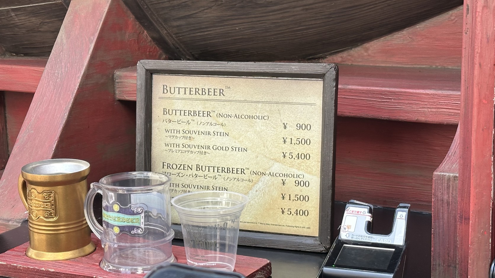 奶油啤酒價格牌：無酒精奶油啤酒與冰沙版皆為一般杯 900 日圓、附紀念馬克杯 1,500 日圓、附金色紀念馬克杯 5,400 日圓，牌子旁擺著金色馬克杯、透明紀念杯與一般塑膠杯