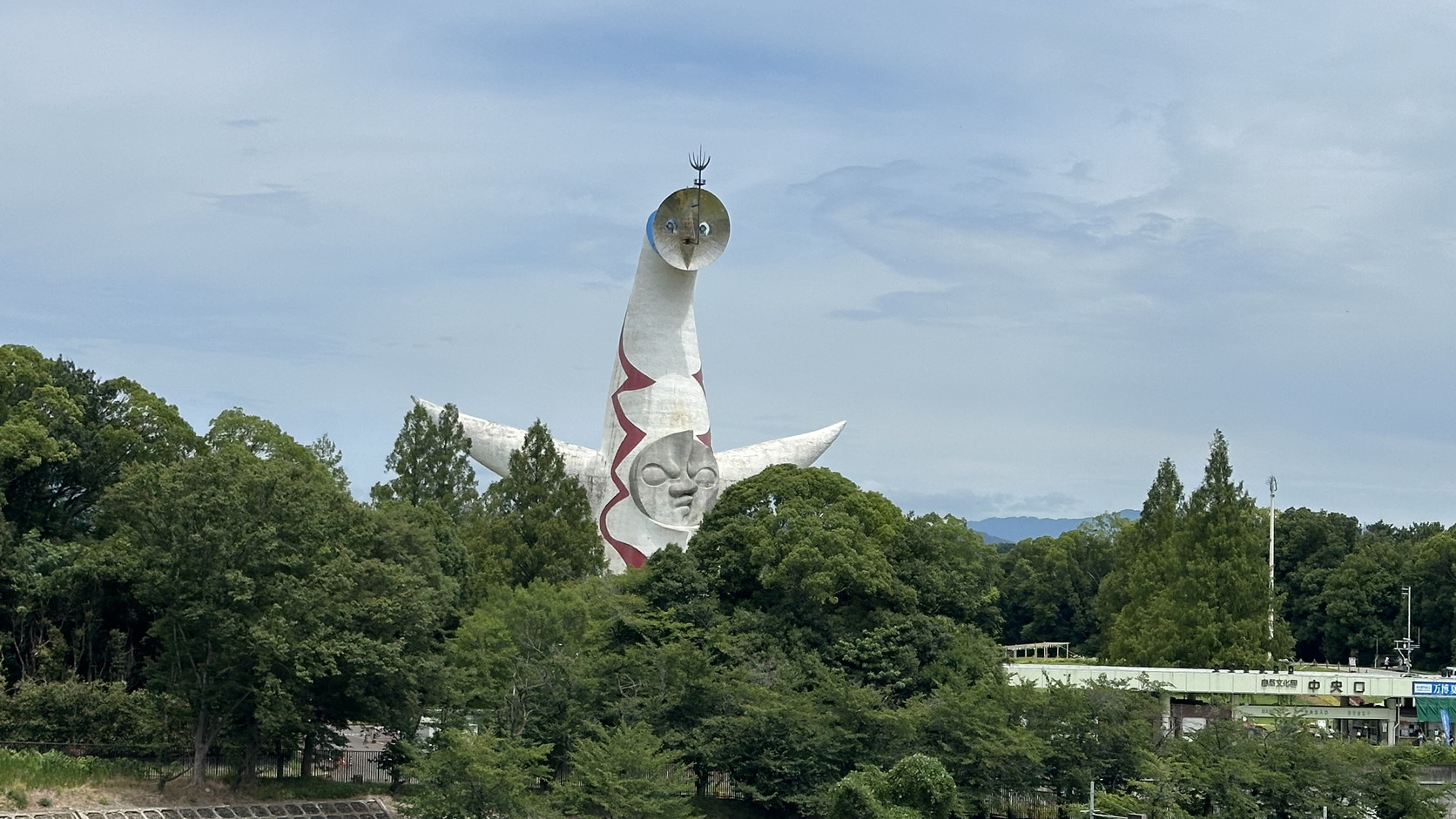 從萬博紀念公園站遠望太陽之塔，塔身從樹林後方升起，塔頂的金色臉清楚可見