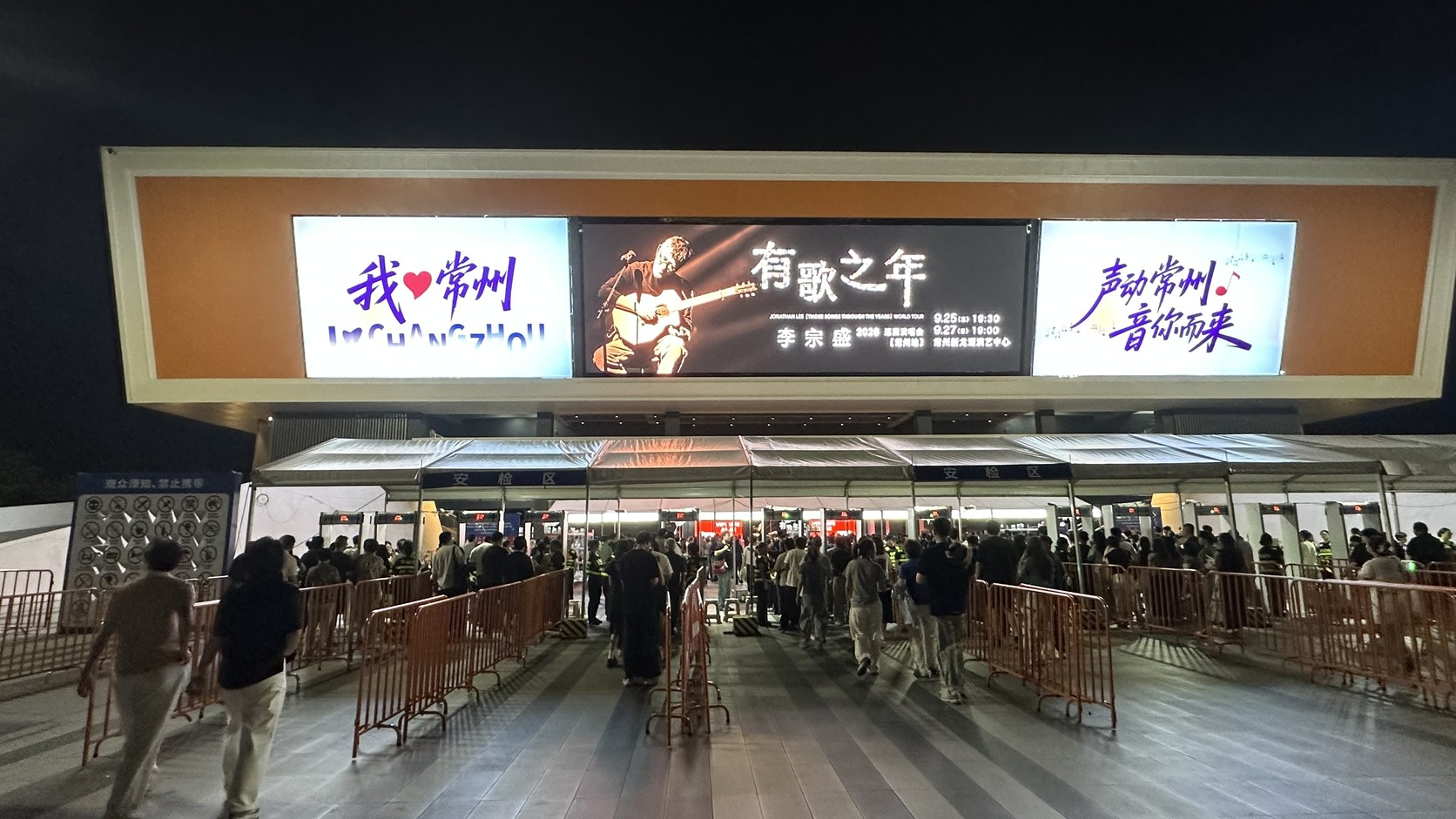 常州新龍湖演藝中心入口：大型螢幕播放《有歌之年》主視覺與場次資訊，安檢區前的欄杆讓進場人潮分流