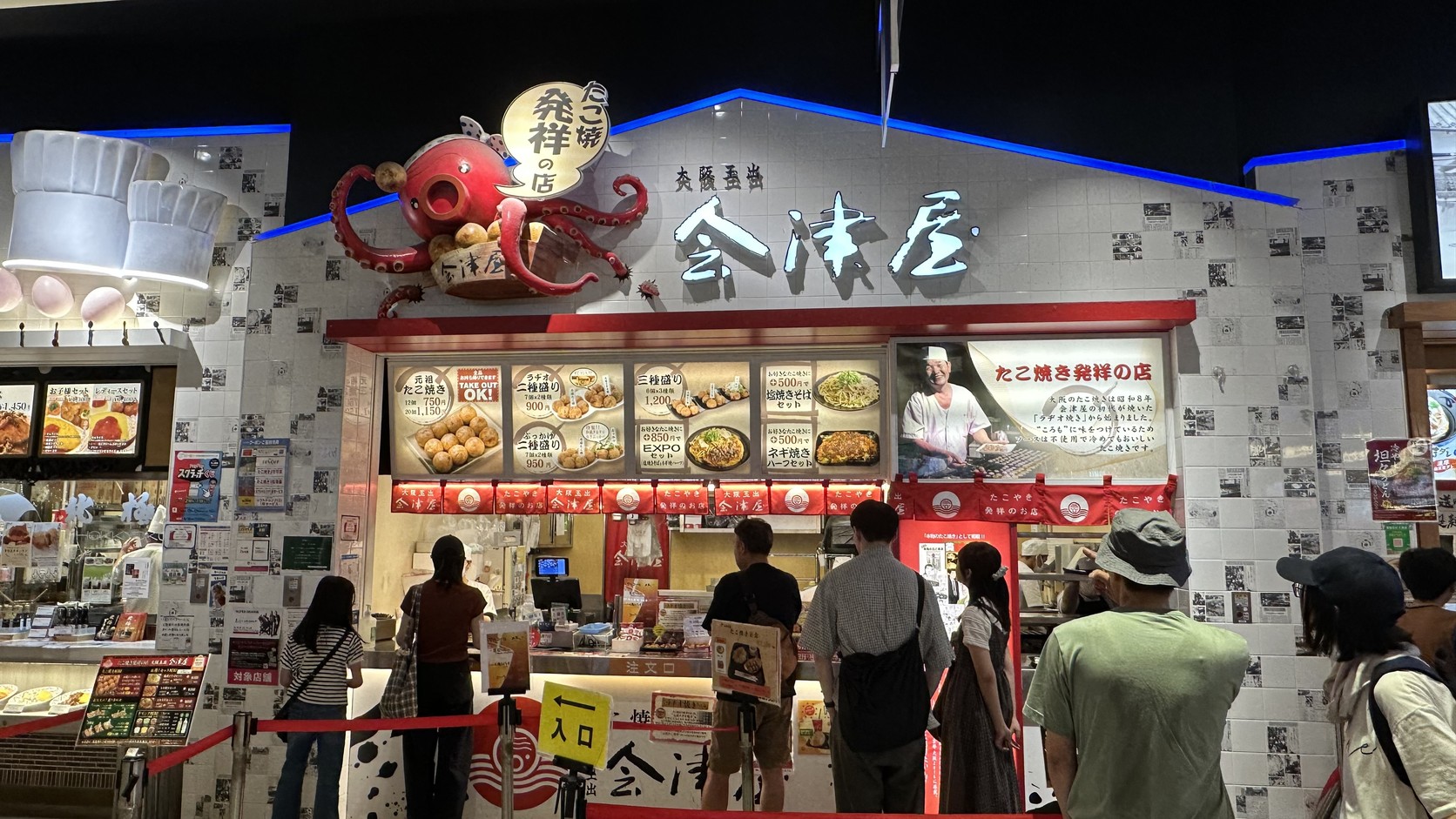 美食廣場裡会津屋的章魚燒店面，招牌寫著「たこ焼き発祥の店」，前方有排隊的客人