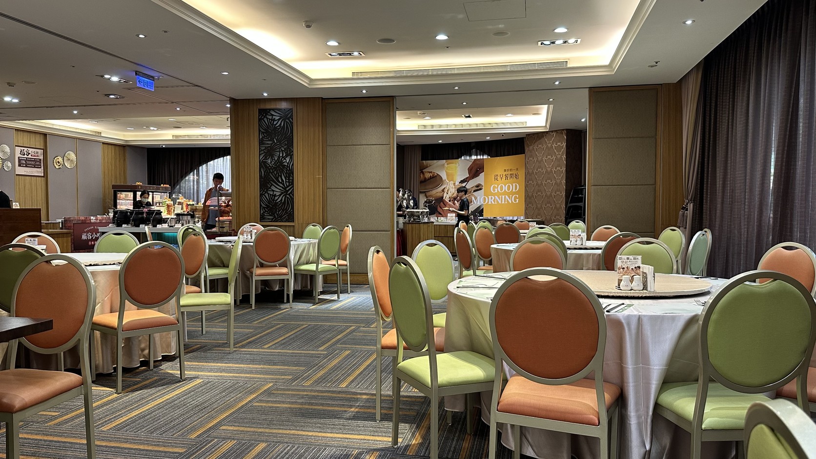 飯店二樓像宴會廳一樣的早餐用餐空間，擺著許多圓桌與綠橘色座椅