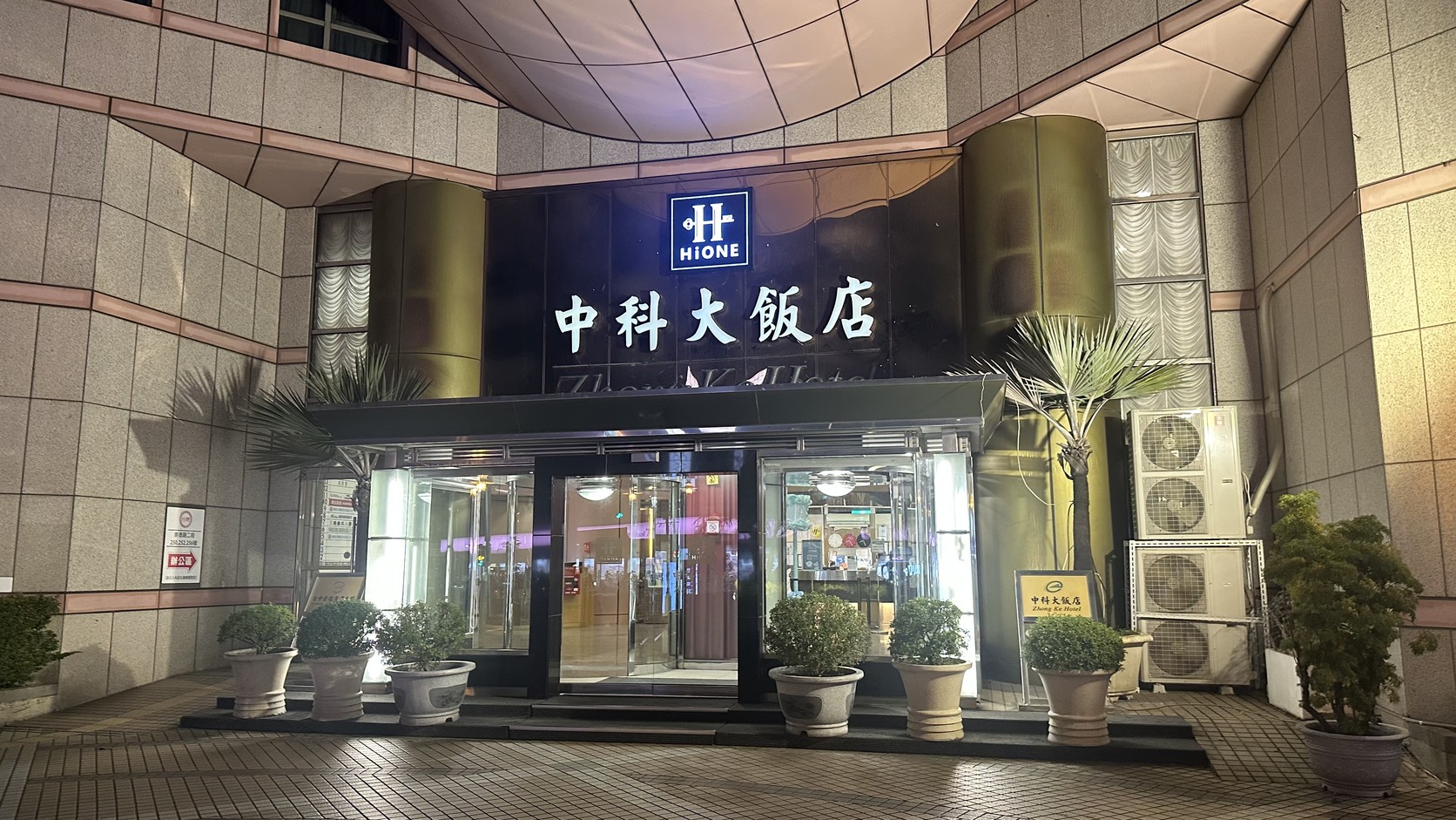 中科大飯店夜間入口：深色招牌上有 HIONE 標誌與「中科大飯店」白字，門前擺著盆栽