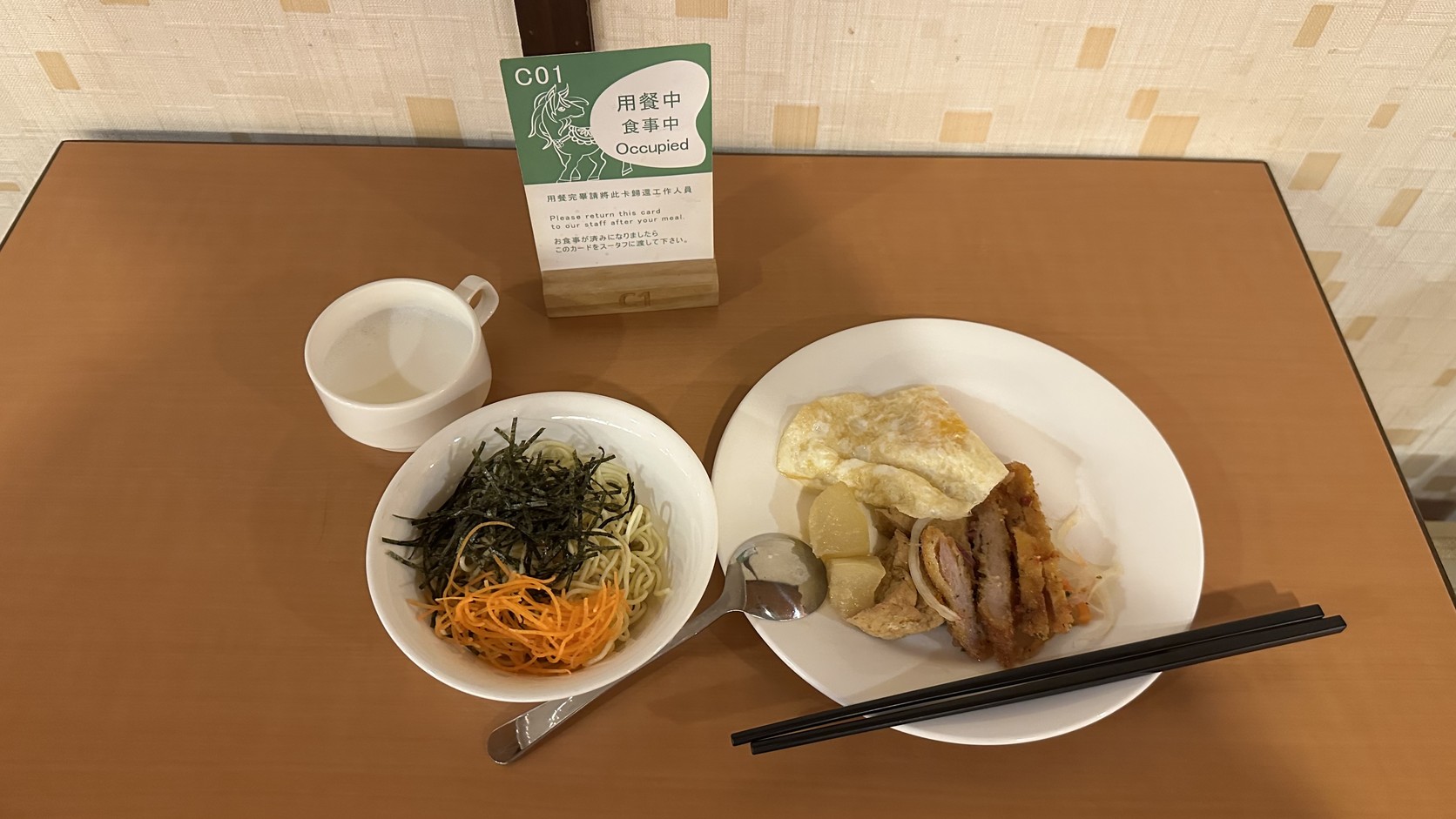 日式冷麵餐盤：細麵上鋪著海苔絲與紅蘿蔔絲，旁邊是煎蛋與肉類料理，桌上放著用餐中桌牌