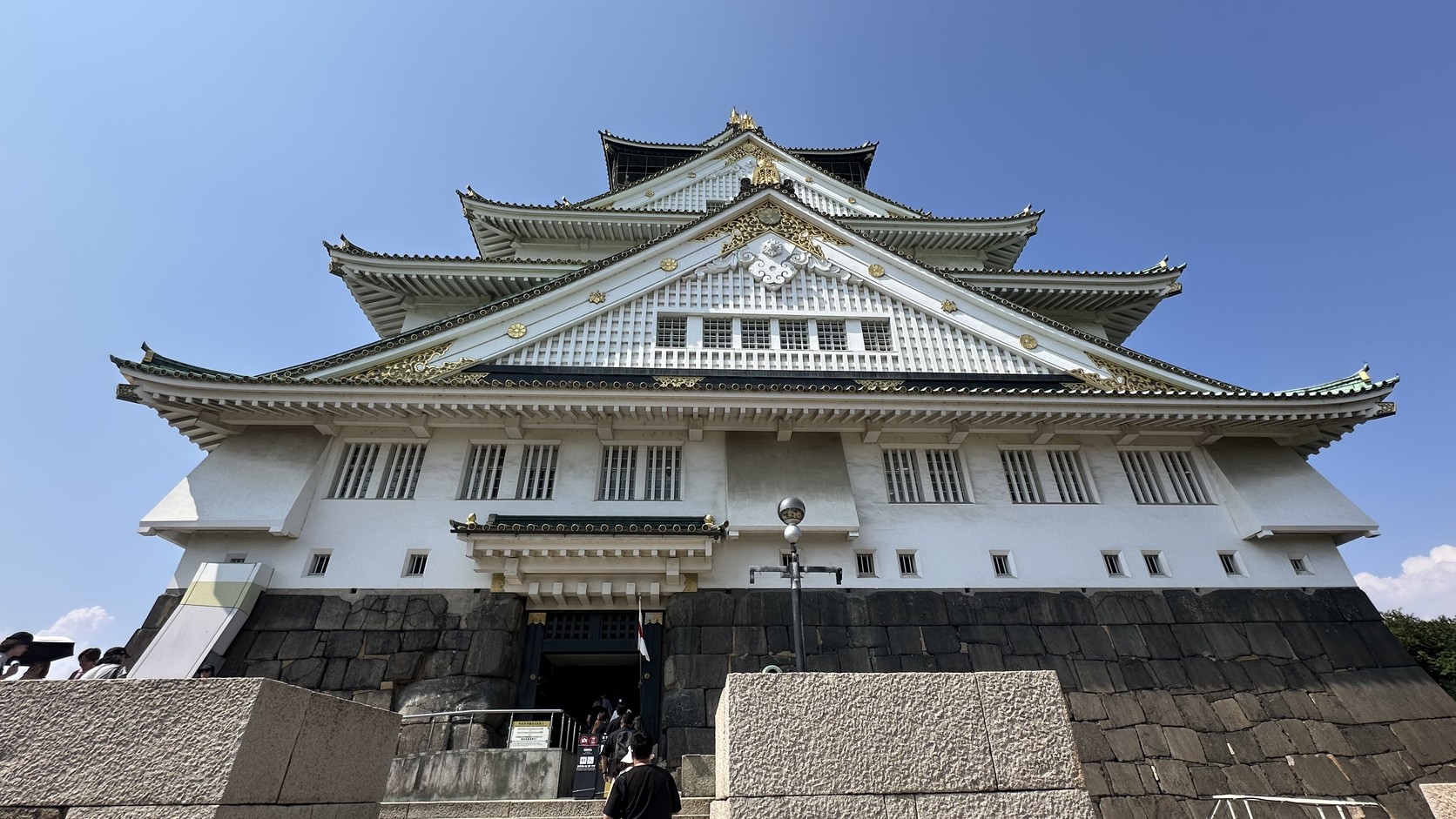 從石垣下仰望大阪城天守閣：白牆、綠瓦屋簷與金色裝飾層層向上，背後是藍天