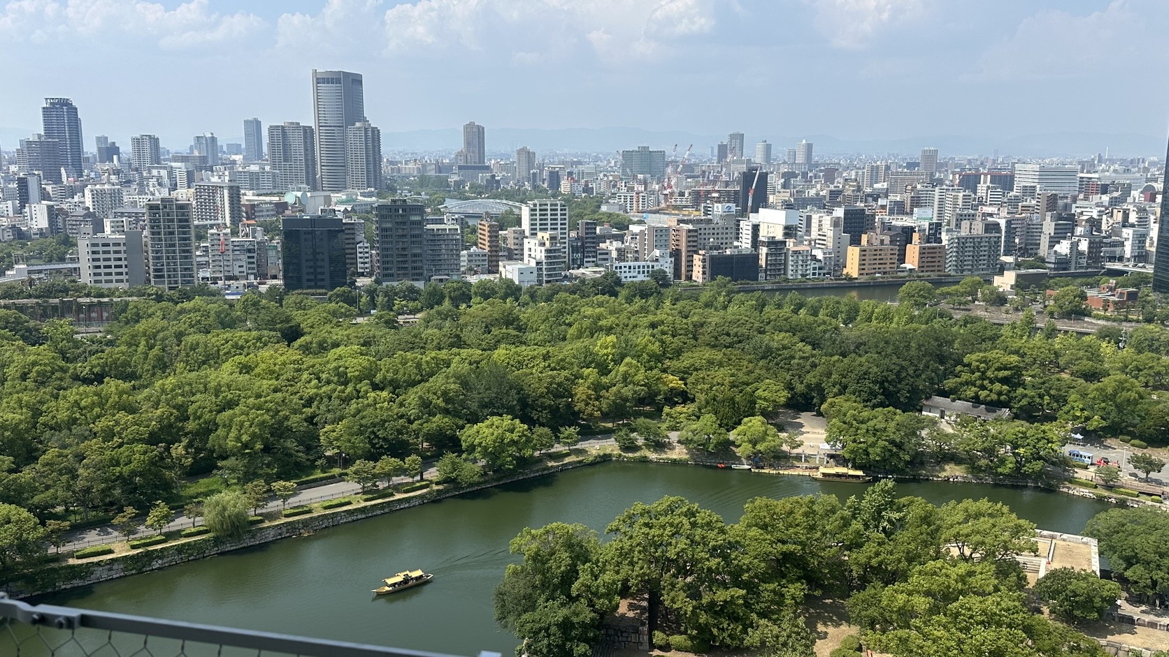 從大阪城天守閣八樓展望台俯瞰護城河與公園綠地，河中有一艘金色的御座船，遠處是大阪市區