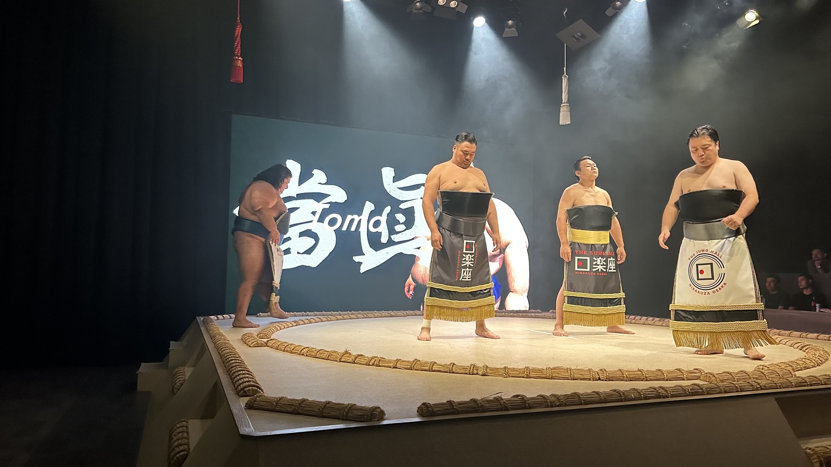 大阪日樂座相撲表演：三位力士身穿化粧廻し登上土俵，背後大型螢幕顯示表演標題
