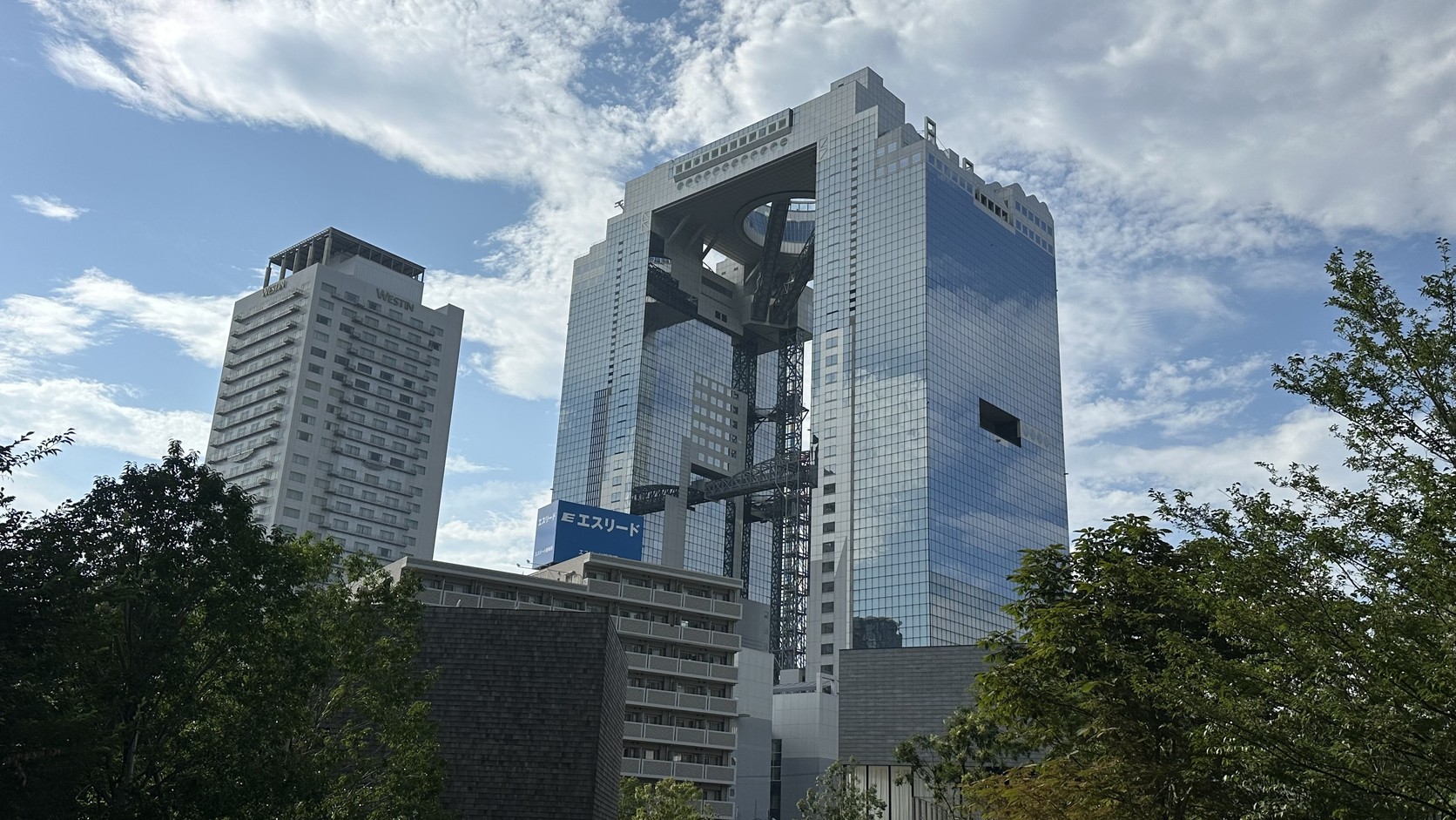 從遠處望向梅田藍天大廈的雙塔，銀灰色玻璃帷幕映著藍天白雲，兩棟高樓在頂端相連