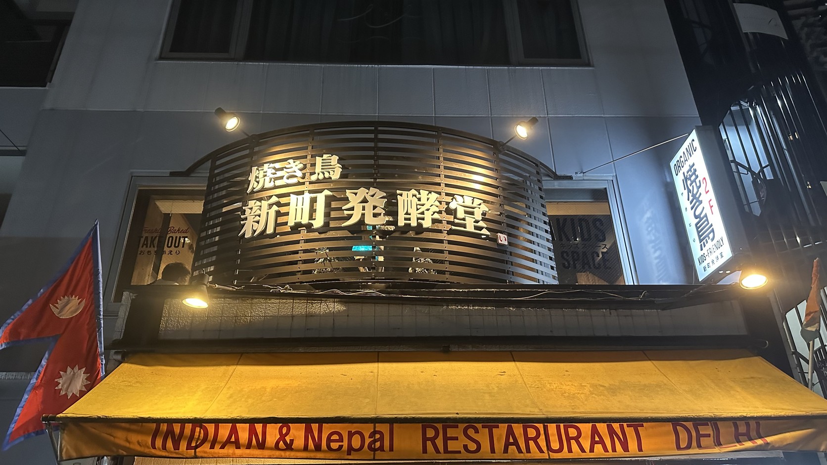 「焼き鳥 新町発酵堂」夜間招牌與二樓直立招牌，樓下是掛著黃色遮陽棚的印度與尼泊爾餐廳