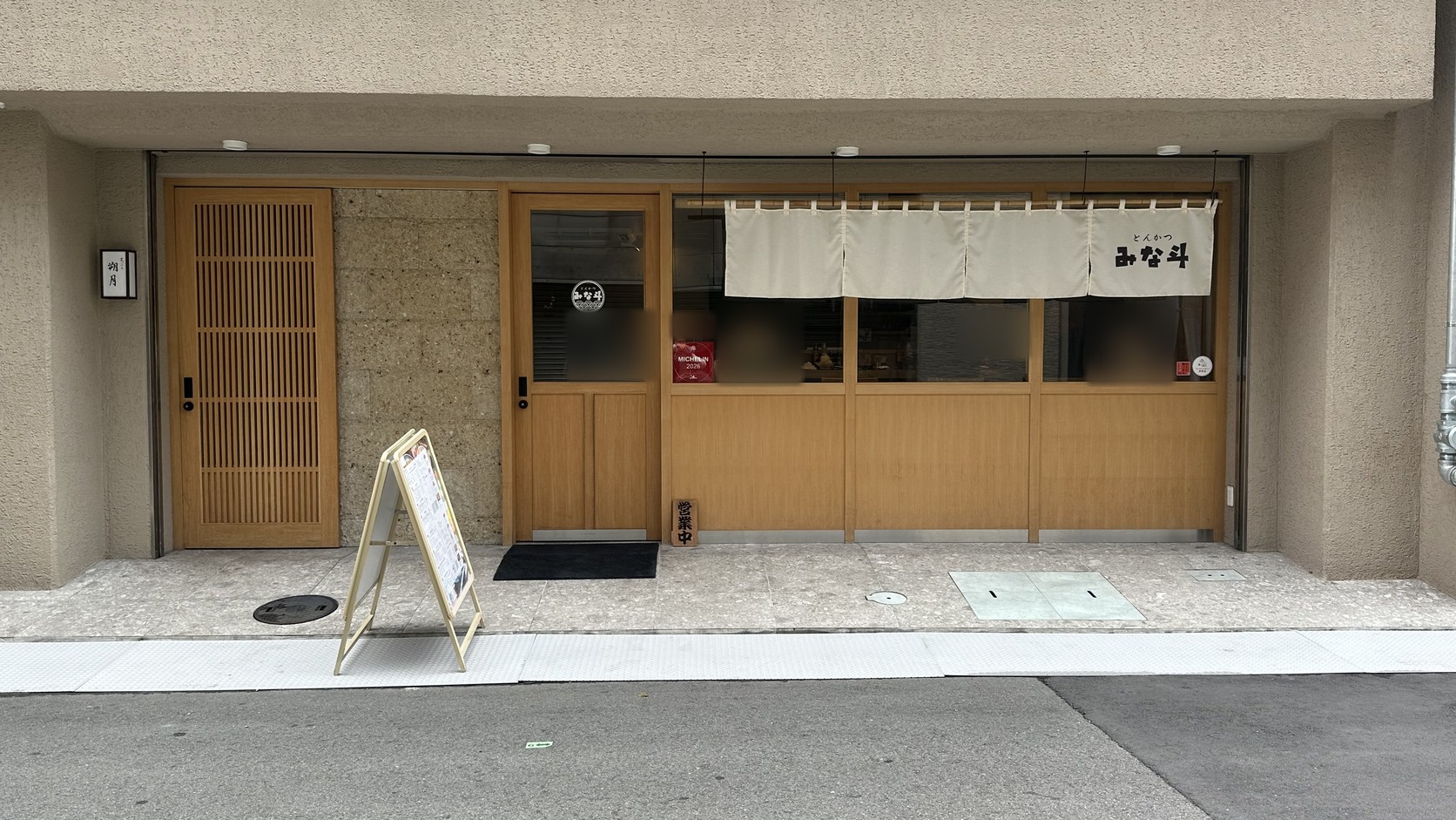 「とんかつ みな斗」店面外觀，白色暖簾上寫著「とんかつ みな斗」，旁邊是木質門窗、入口與立牌菜單，窗內用餐客人已做模糊處理