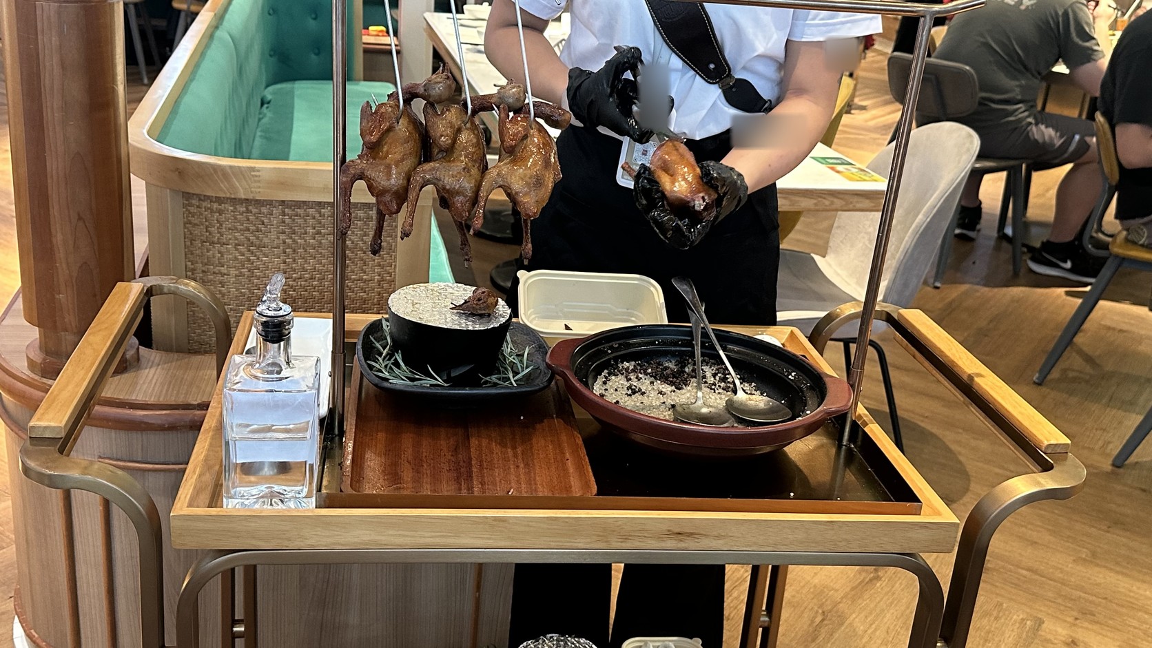 店員推著餐車到桌邊並剪切乳鴿的實拍，餐車上懸掛著數隻乳鴿，店員的手臂刺青與工作證已模糊