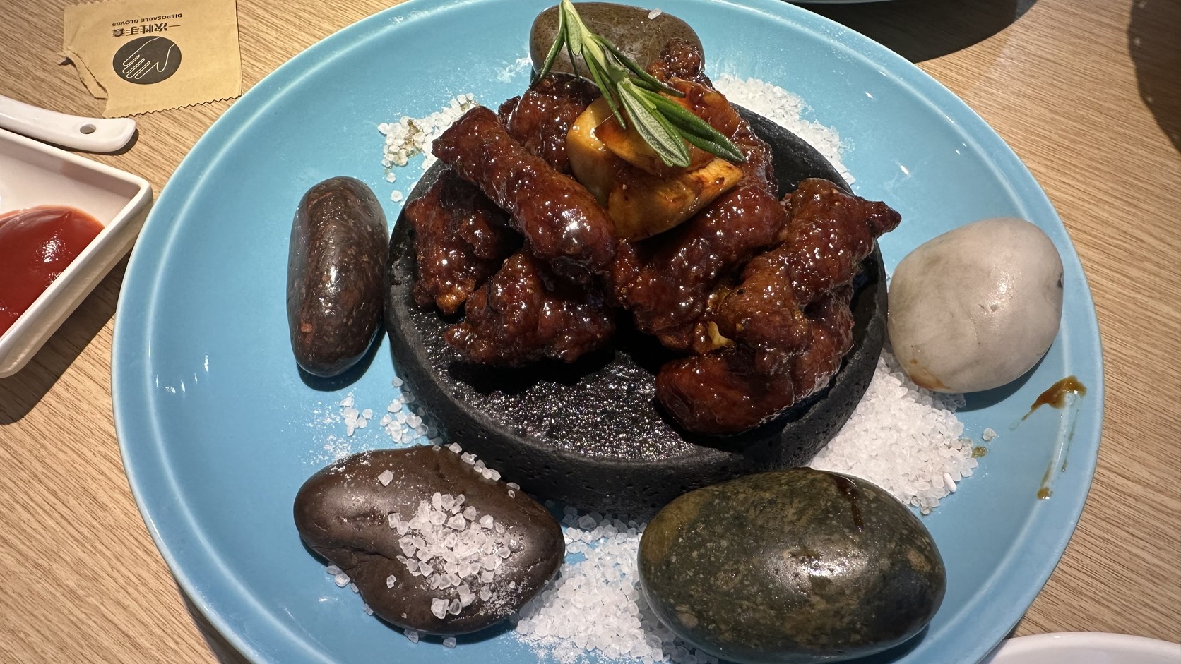 金蒜脆皮牛肉粒實拍，裹著深色醬汁的牛肉粒放在黑色石板上，周圍有鵝卵石、白色粗顆粒與迷迭香