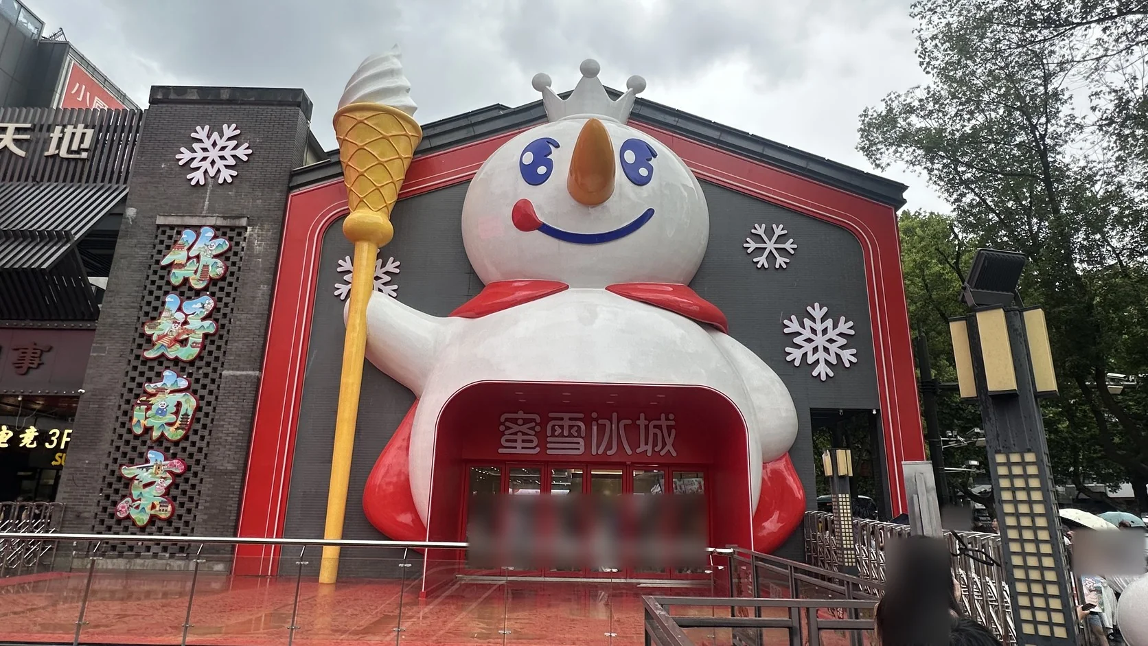 南京夫子廟蜜雪冰城旗艦店外觀：戴皇冠、舉冰淇淋權杖的巨大雪王成為入口，旁邊是直式「你好南京」招牌（入口人潮已模糊）