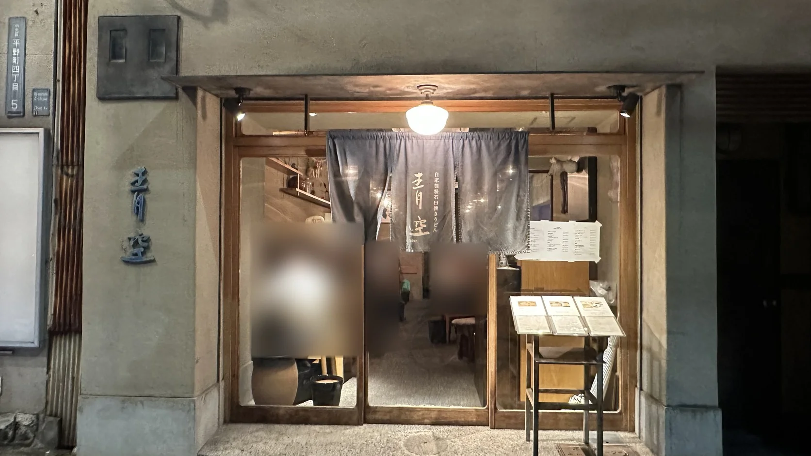 青空blue店面外觀：藍色暖簾上寫著「自家製粉石臼挽きうどん」，木框玻璃門與門口菜單架，窗內用餐客人已做模糊處理