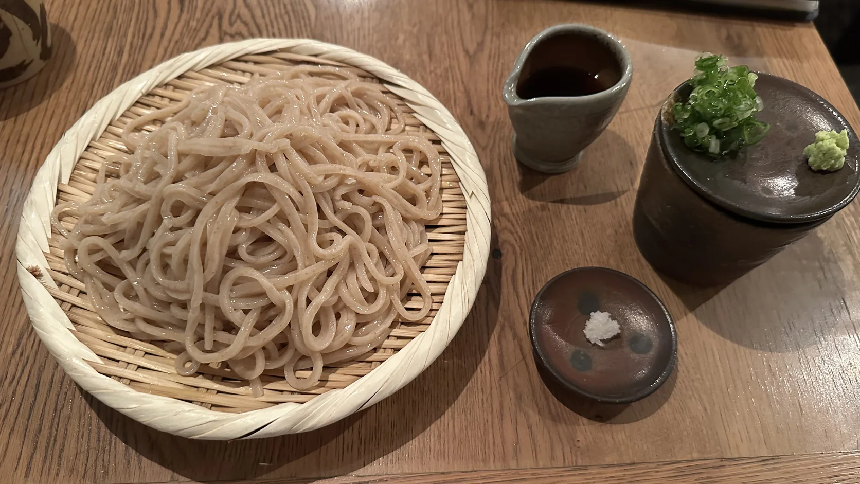 青空blue粗磨冷烏龍麵：赤褐色麵條盤成圈放在有縫隙的圓形竹簾上，旁邊是小壺沾汁、蓋上放著蔥花與山葵的大杯和一碟鹽