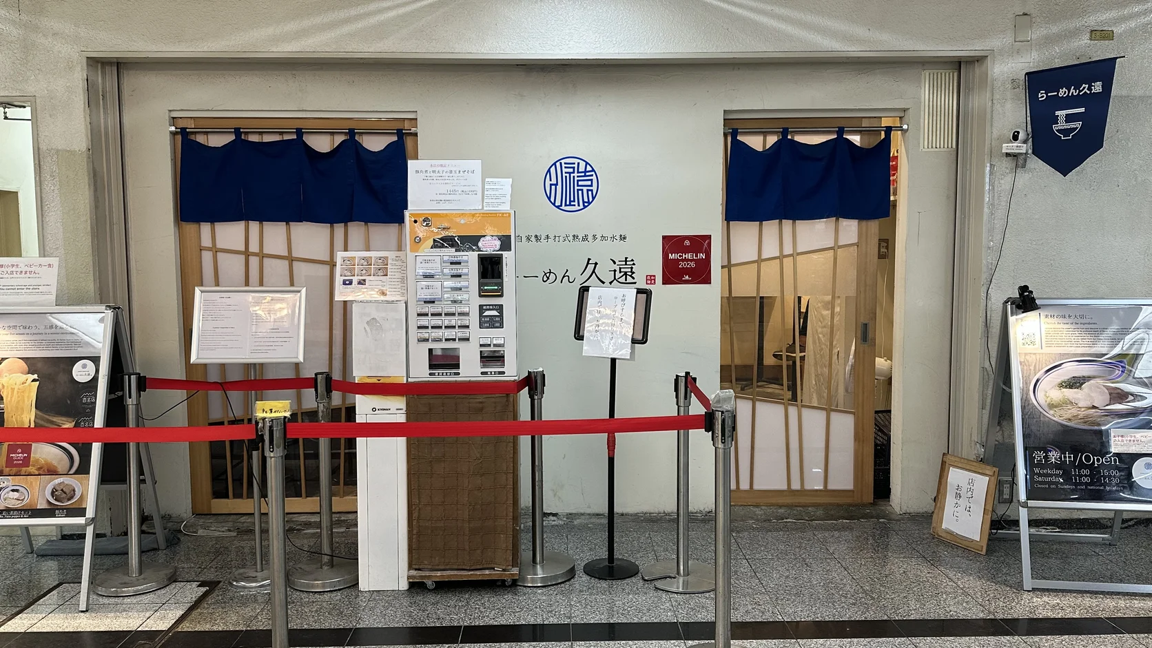 らーめん久遠店面：白牆上有店名與紅色 MICHELIN 2026 標誌，門口設有售票機，前方拉著紅色隔離帶
