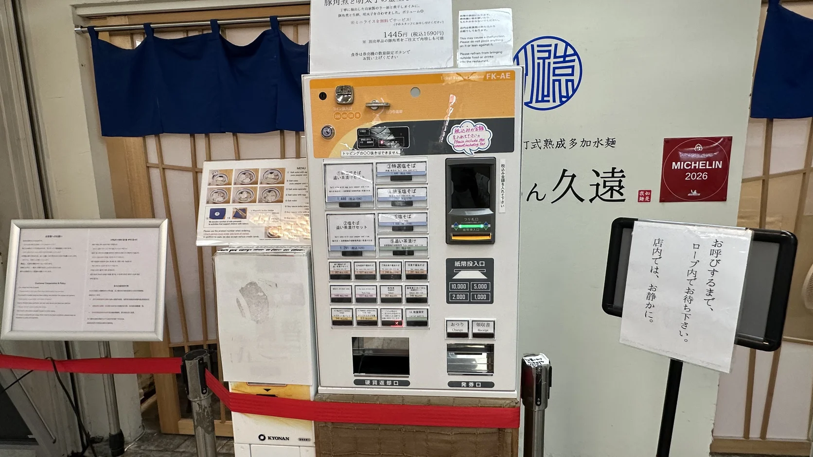 店門口的售票機，上方有餐點按鍵與投幣口，旁邊貼著請在線內等候的告示