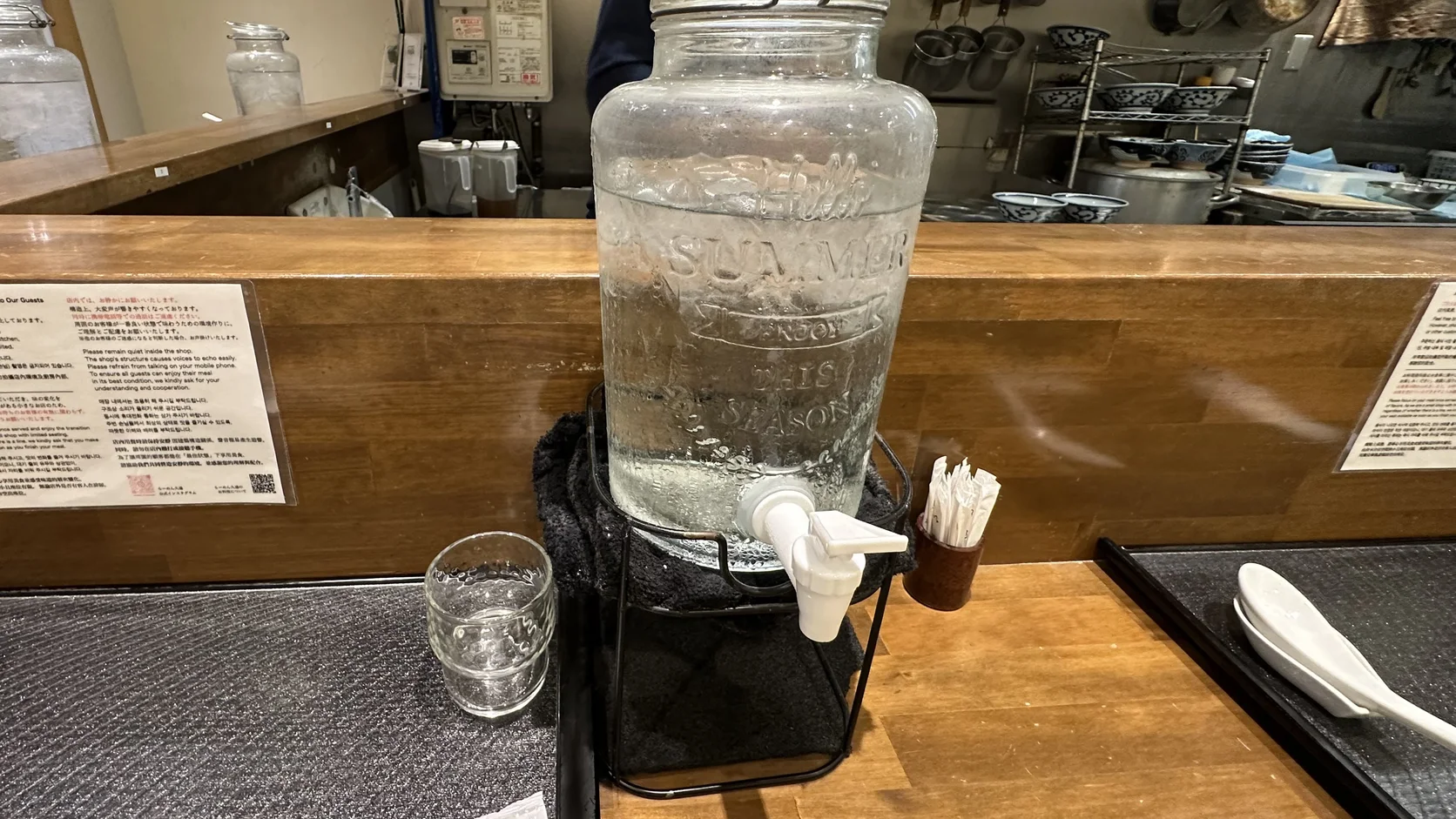 吧檯上的大型玻璃飲水罐，下方有白色水龍頭，旁邊放著一只玻璃杯