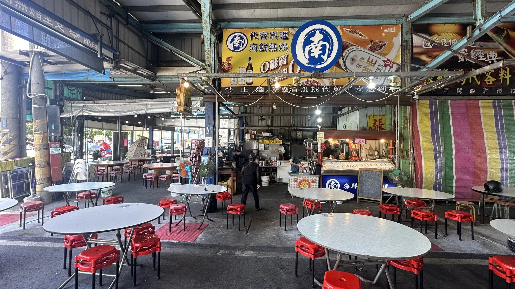 南董店內全景：半開放式空間裡擺著圓桌與紅色塑膠凳，前方是料理區與海鮮展示櫃，上方掛著「代客料理、海鮮熱炒」看板