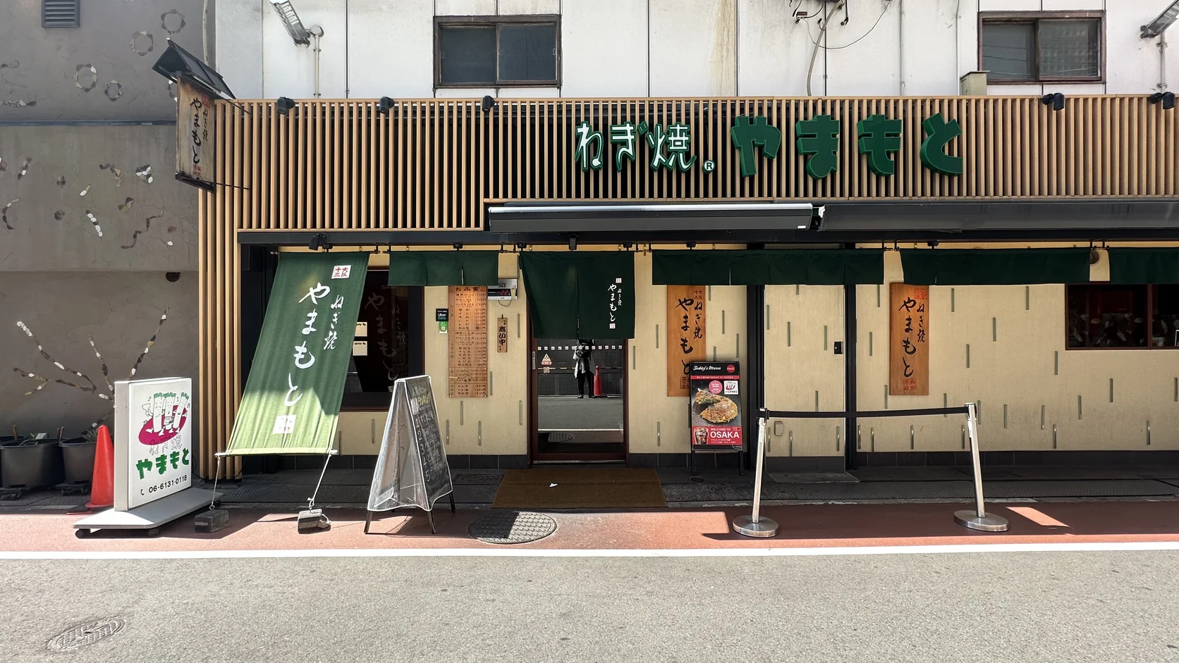 山本蔥燒梅田EST店的店面外觀，木質格柵上方是綠色「ねぎ焼 やまもと」招牌，門口垂著綠色暖簾與布旗