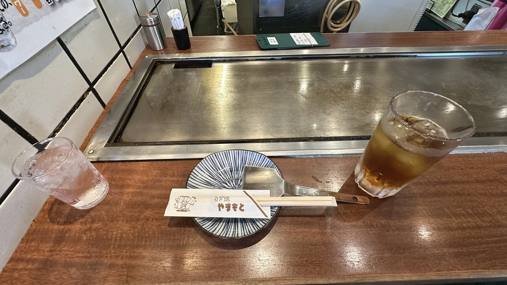 吧台前嵌著一塊長方形鐵板，旁邊是小盤、小鏟子與印有山本蔥燒標誌的竹筷，還有兩杯飲料