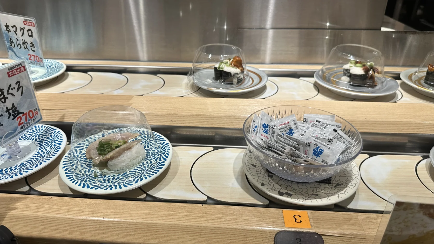 迴轉輸送帶上的壽司盤加了透明保鮮蓋，旁邊有一碗小包裝調味料