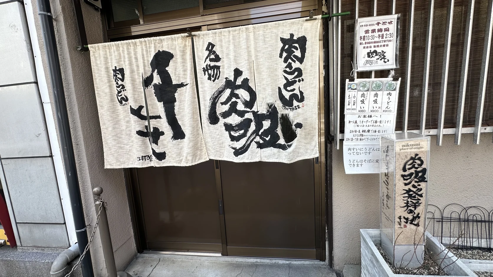 千とせ本店門口的米白色暖簾，寫著「千とせ」「名物 肉吸」「肉うどん」，右側是「肉吸い発祥の地」石碑與門旁公告