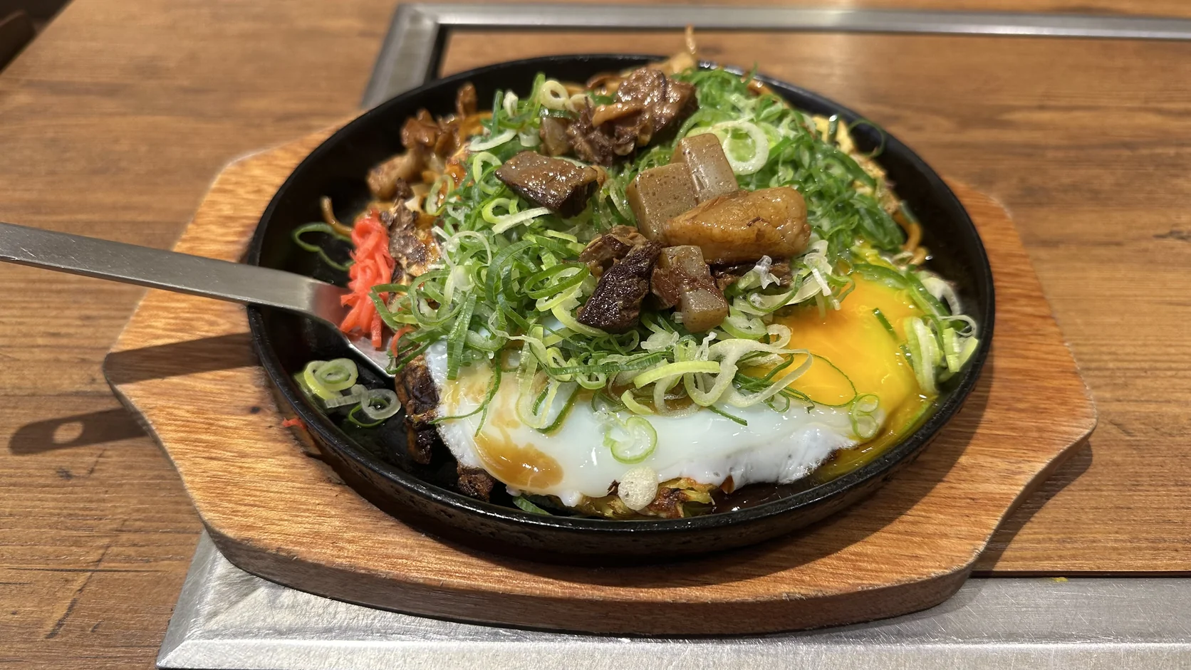 黑色鐵盤上的青蔥牛筋大阪燒炒麵套餐，鋪滿青蔥，旁邊有牛筋、蒟蒻、半熟煎蛋、紅薑與一把小鏟子