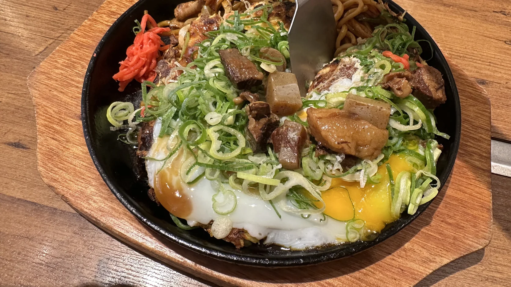 小鏟子切開餐點後的特寫，半熟蛋黃流出，周圍是青蔥、牛筋、蒟蒻與炒麵