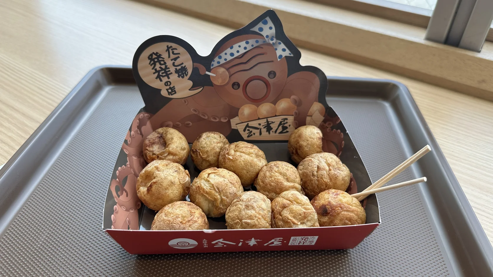 一盒 12 顆會津屋元祖章魚燒，裝在印有章魚吉祥物的紙盒裡，表面沒有淋醬，旁邊放著兩支牙籤